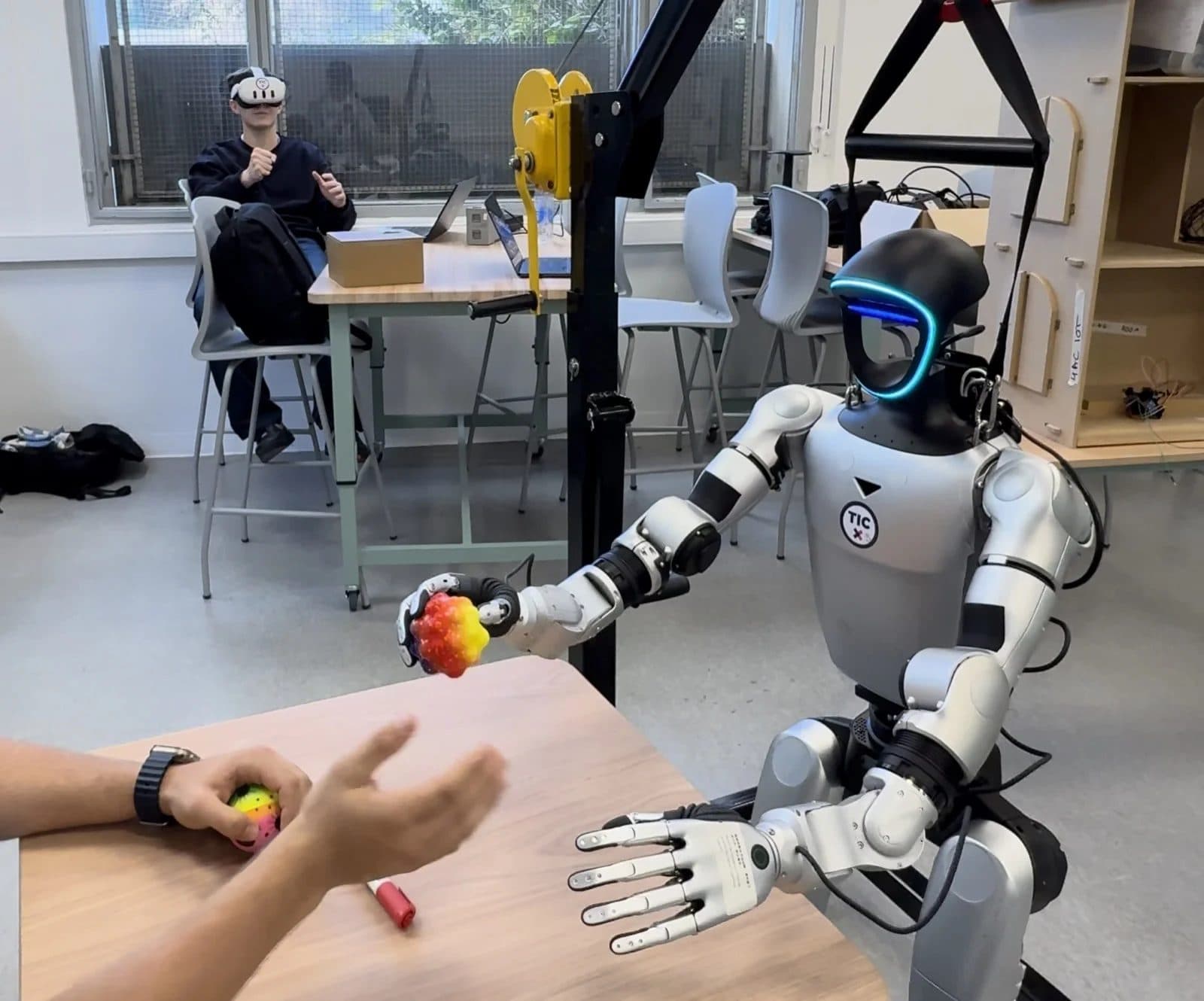 The G1 robot's hand holding a ball while an operator wearing a VR headset controls it in the background